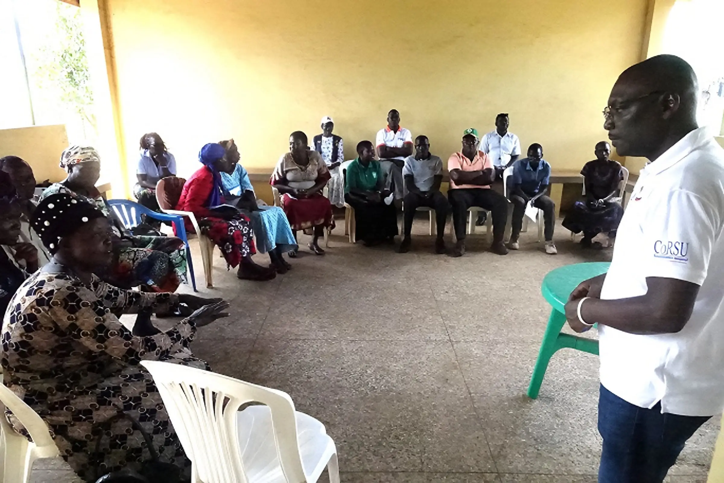 A man in a CoRSU polo shirt speaks to community members seated around a hall.