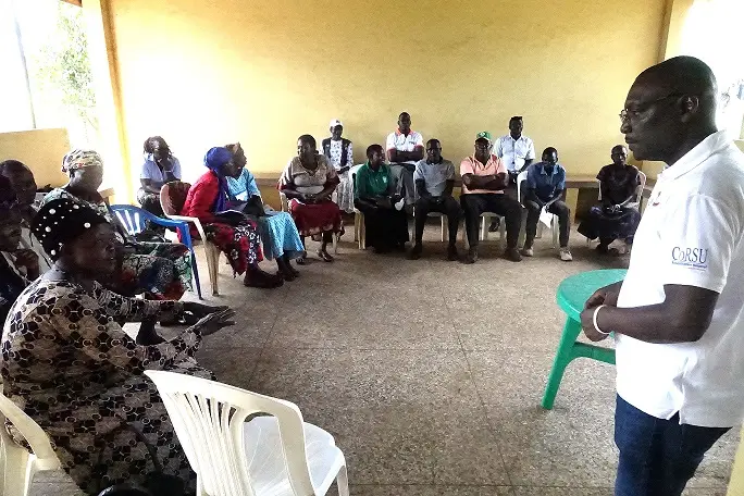 A man in a CoRSU polo shirt speaks to community members seated around a hall.
