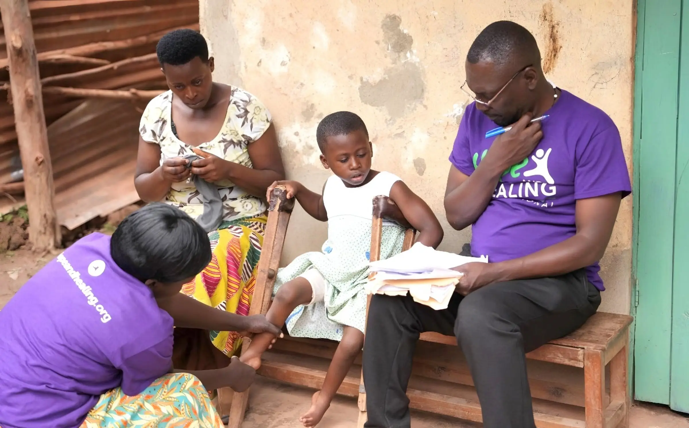 Two staff members in purple T-shirts sit with a mother and her young daughter outside their home, going through papers together.