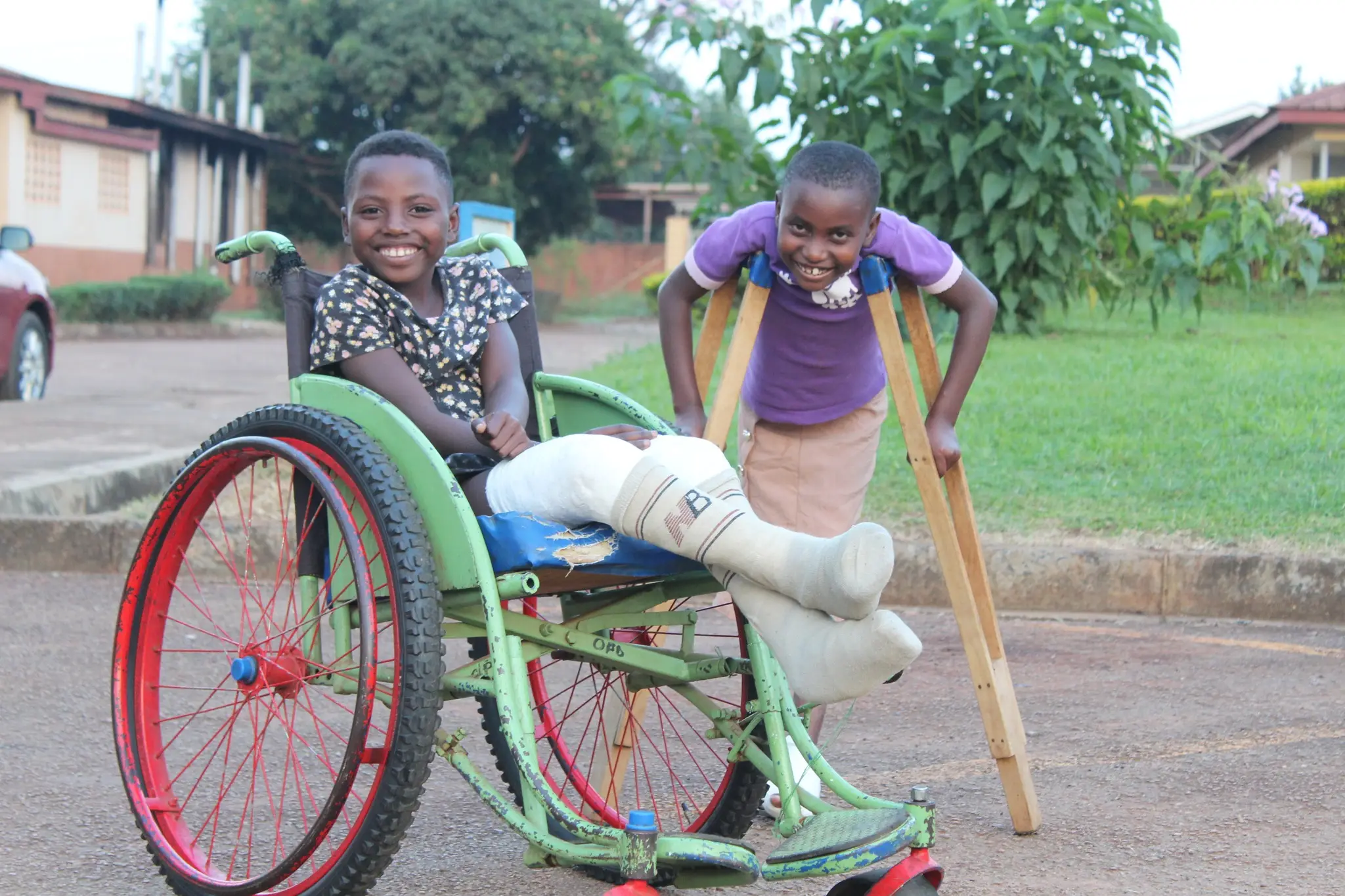 A laughing child in a green wheelchair beside another child on wooden crutches, outside the hospital.