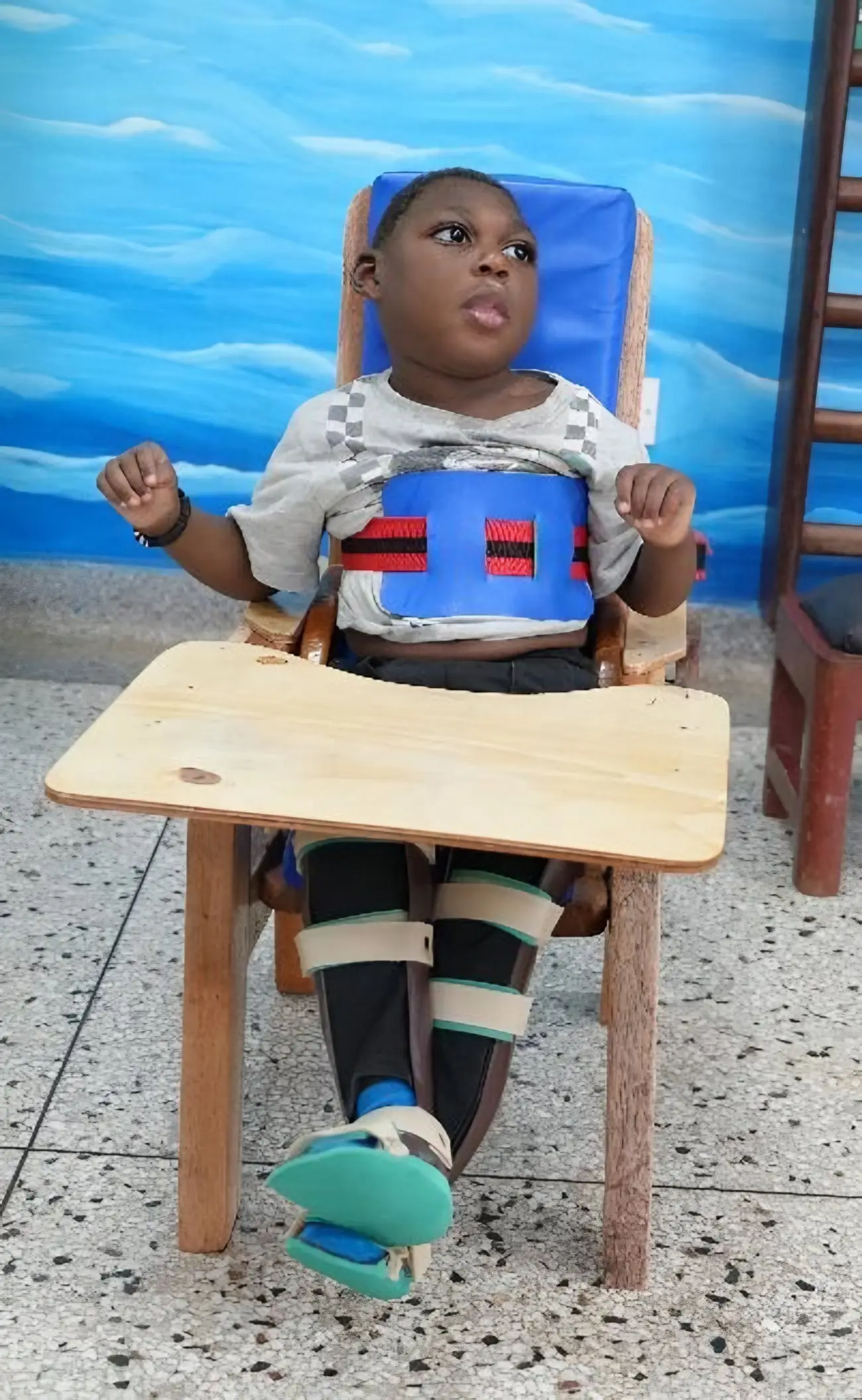 A young boy sits in a supportive therapy chair with a tray, his feet in braces, in front of a painted blue wall.