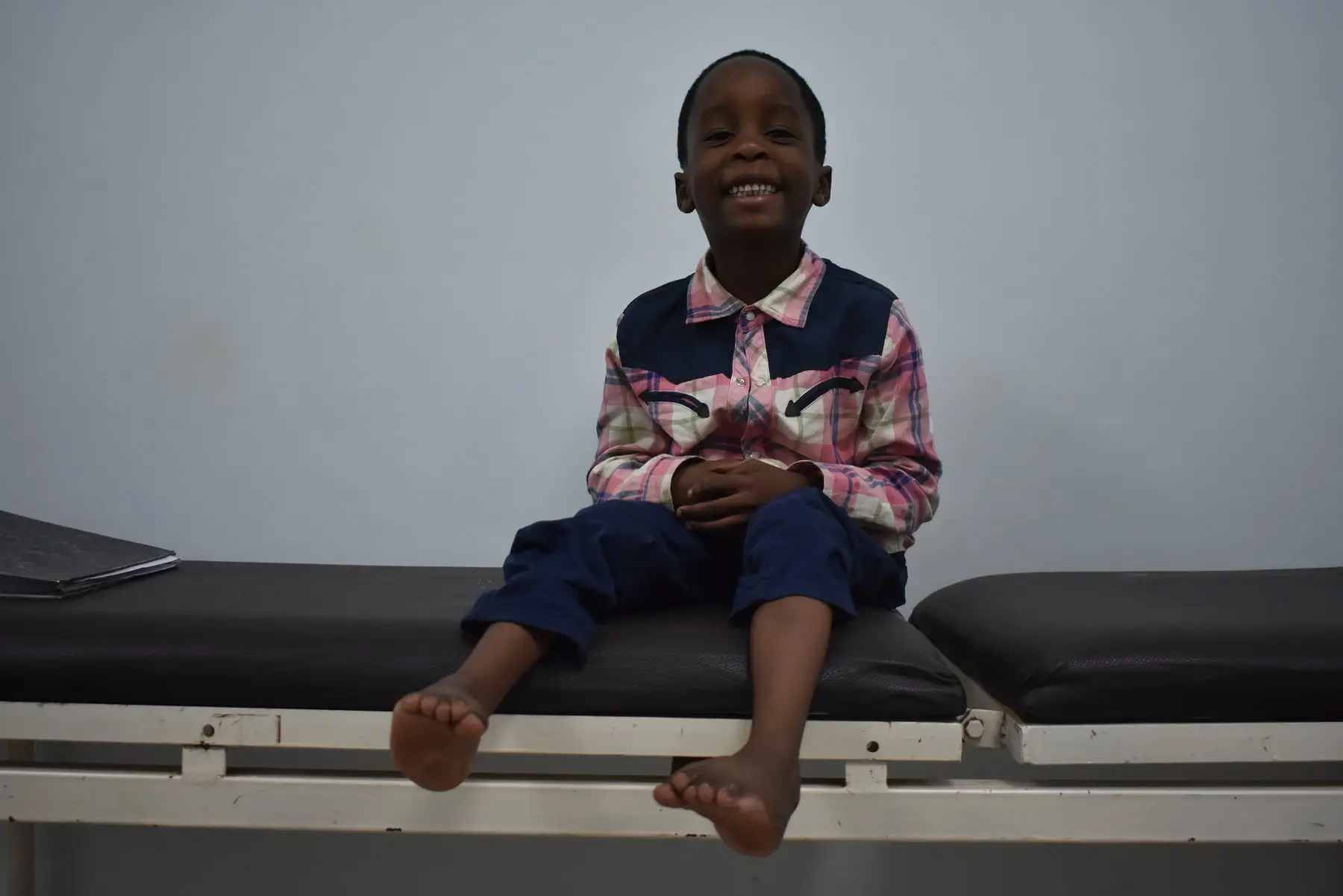 Kimra, a young boy in a pink checked shirt and jeans, smiles as he sits on a treatment couch.