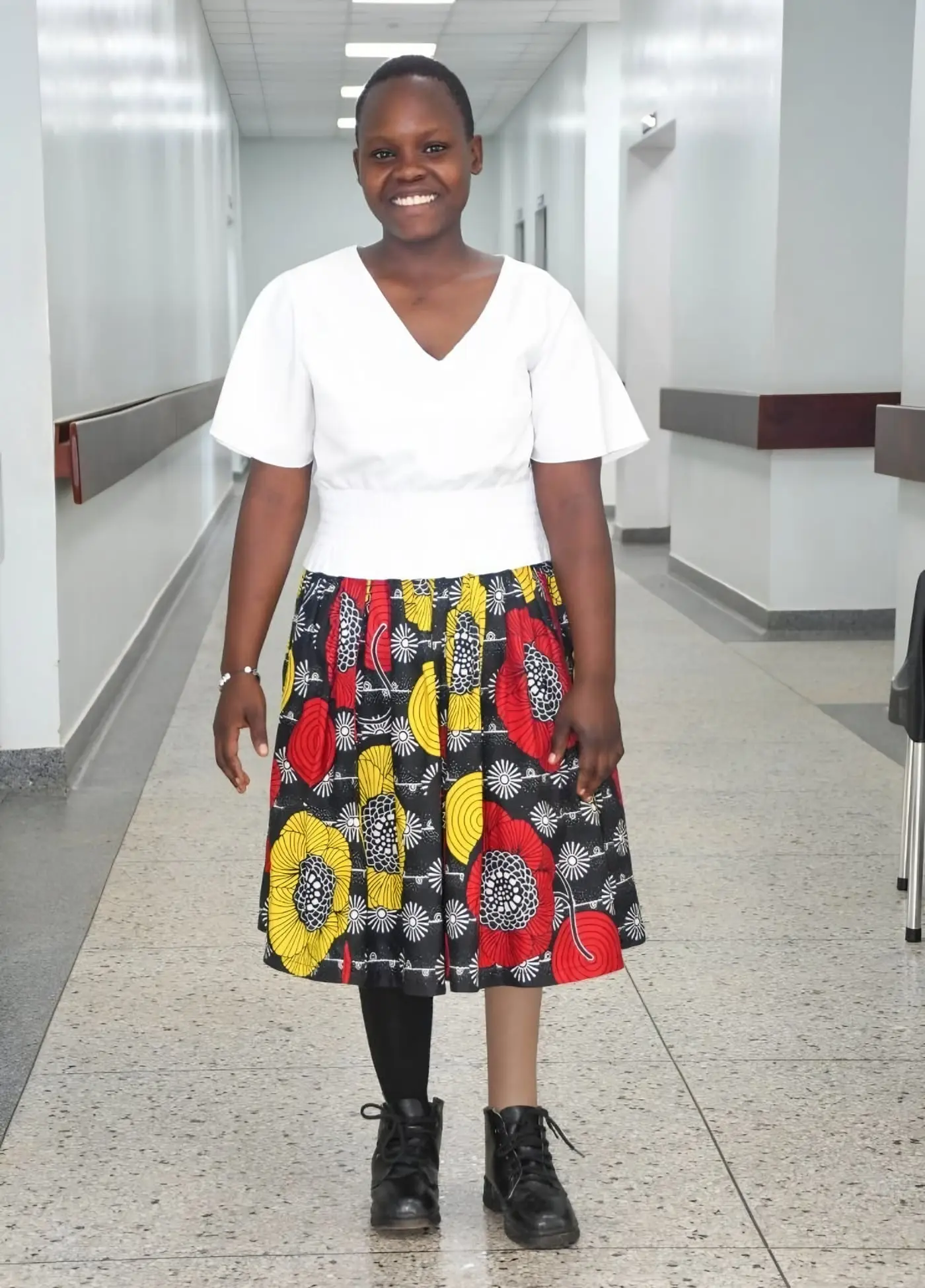 Lilian, a teenage girl in a white top and a bright patterned skirt, smiles as she stands in a hospital corridor on her new prosthesis.