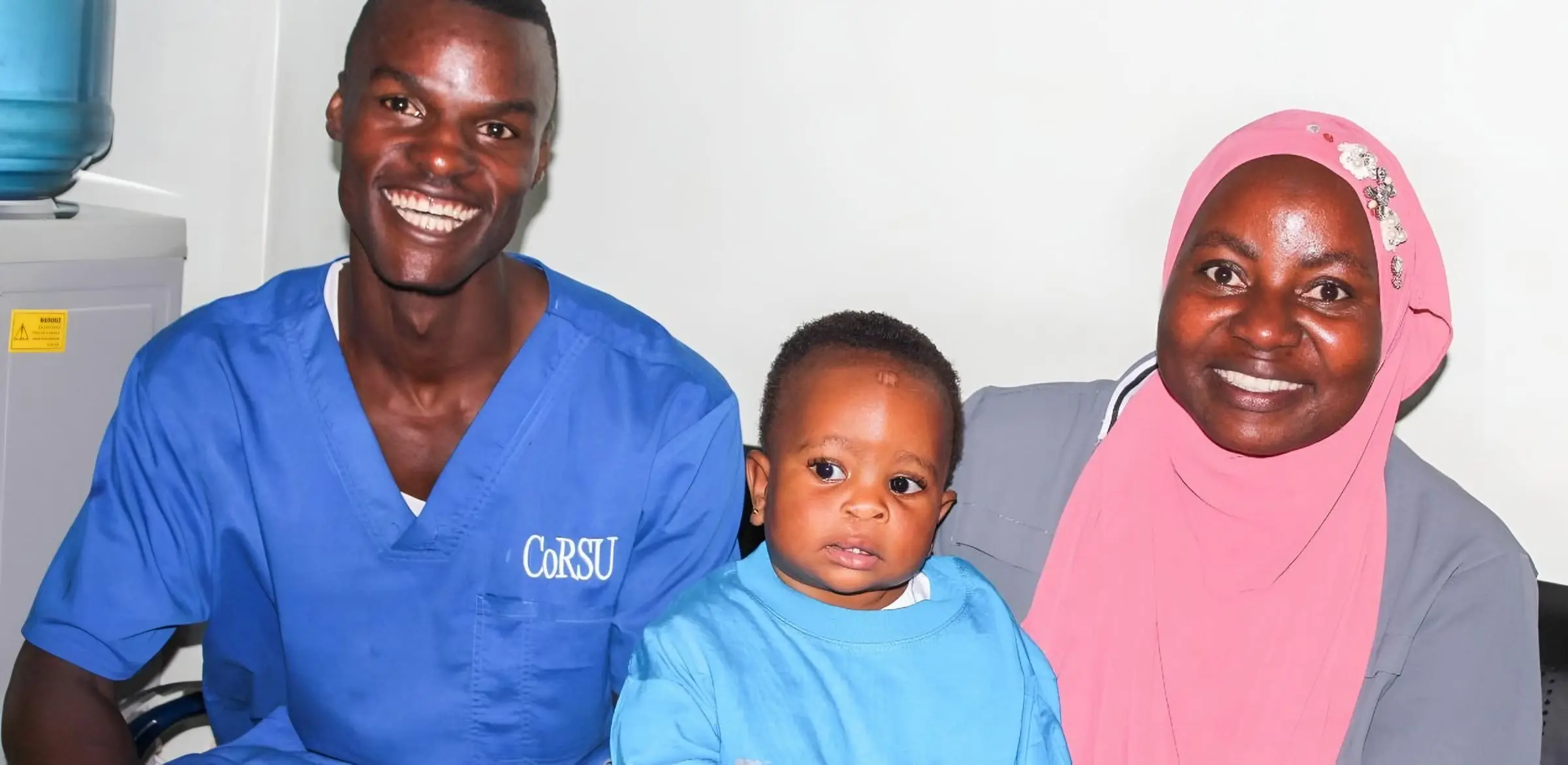 Dr Sadam, in blue CoRSU scrubs, smiles beside Limra and her mother, who wears a pink headscarf.