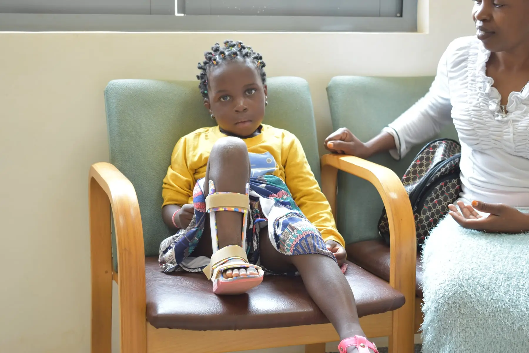 Mercy, a small girl in a yellow top with an orthosis on her right leg, sits in a chair beside her mother.