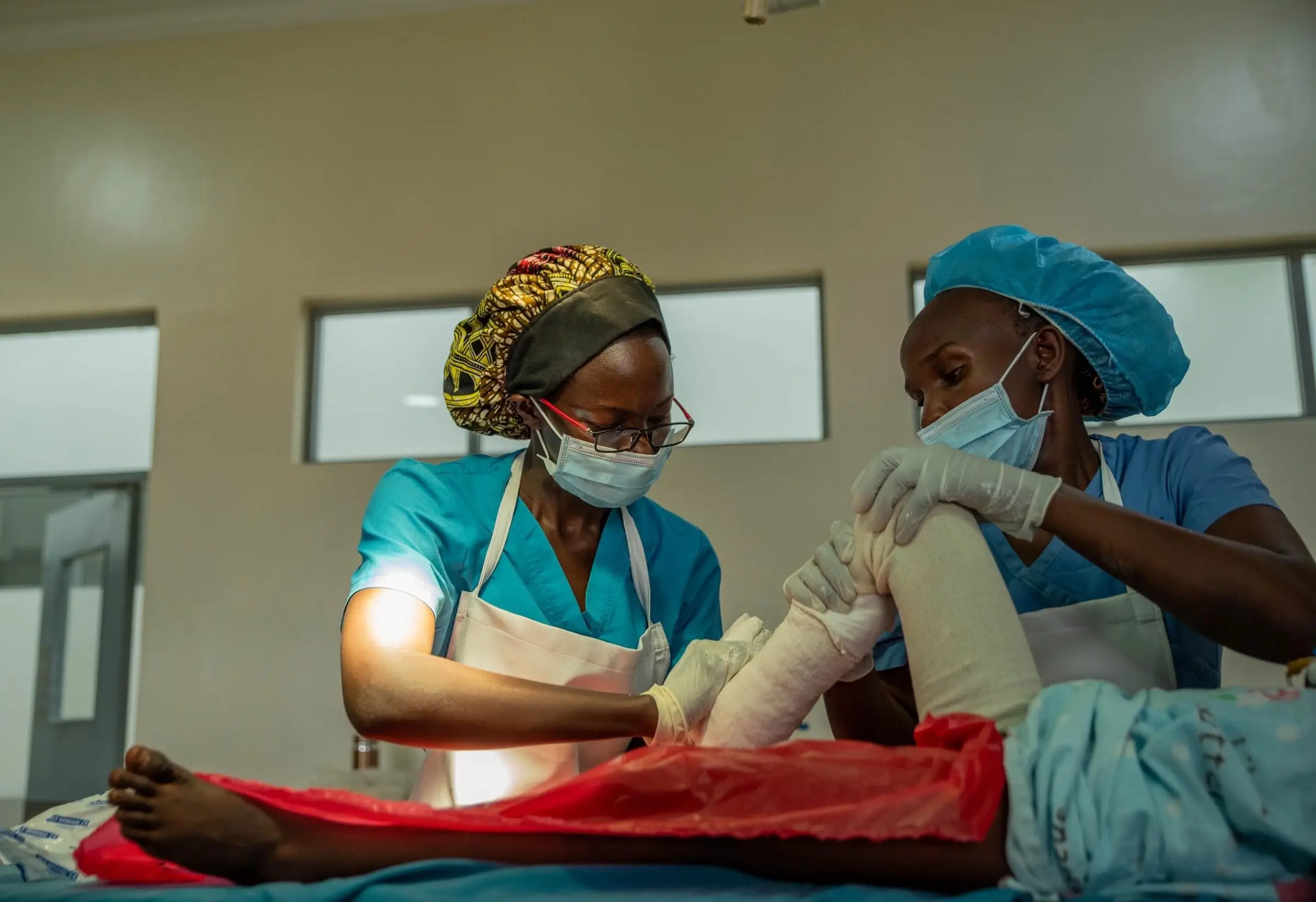 Two clinicians in scrubs, caps and masks apply a cast to a child's leg.