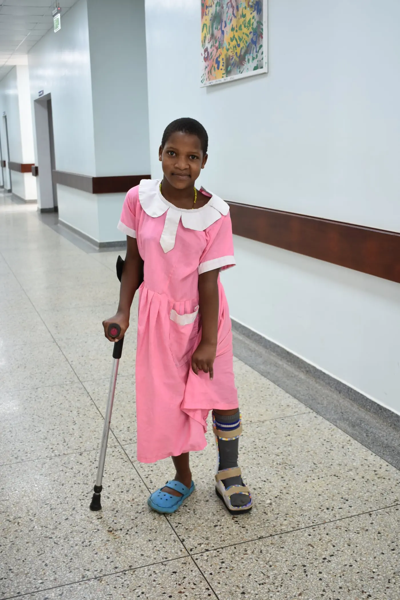 Praise, a girl in a pink dress, stands in a hospital corridor with a crutch and an orthosis on her left leg.