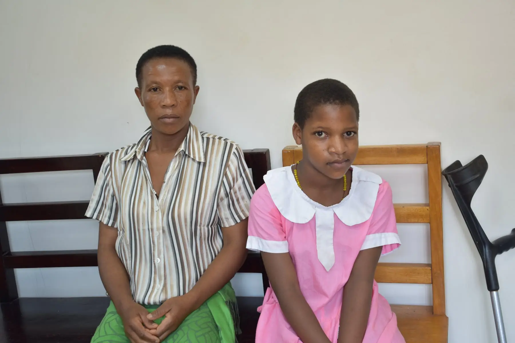 Praise sits on a bench beside her mother, with a crutch resting behind them.