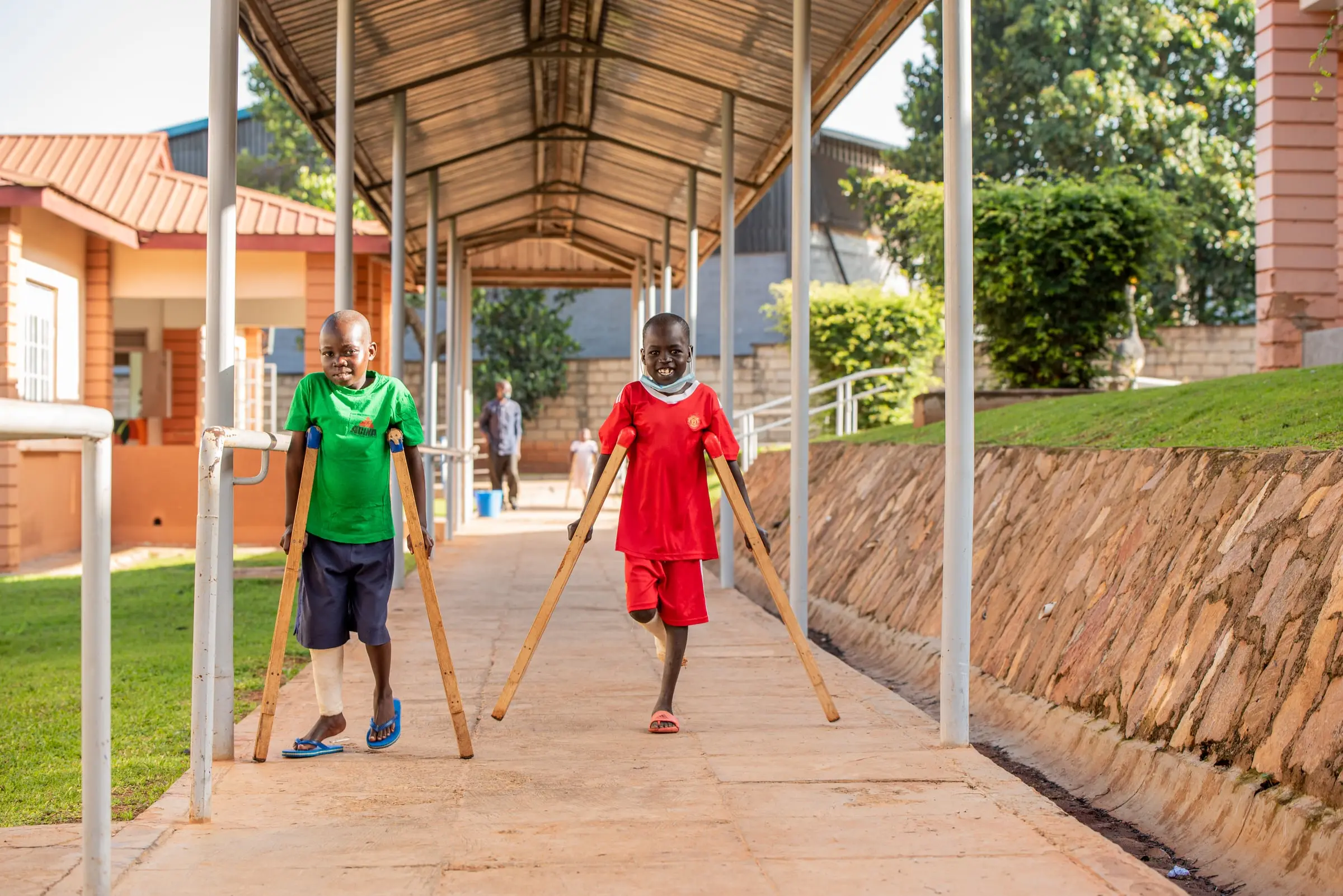 Two boys on crutches, one in green and one in red, walk along a covered walkway in the hospital grounds.