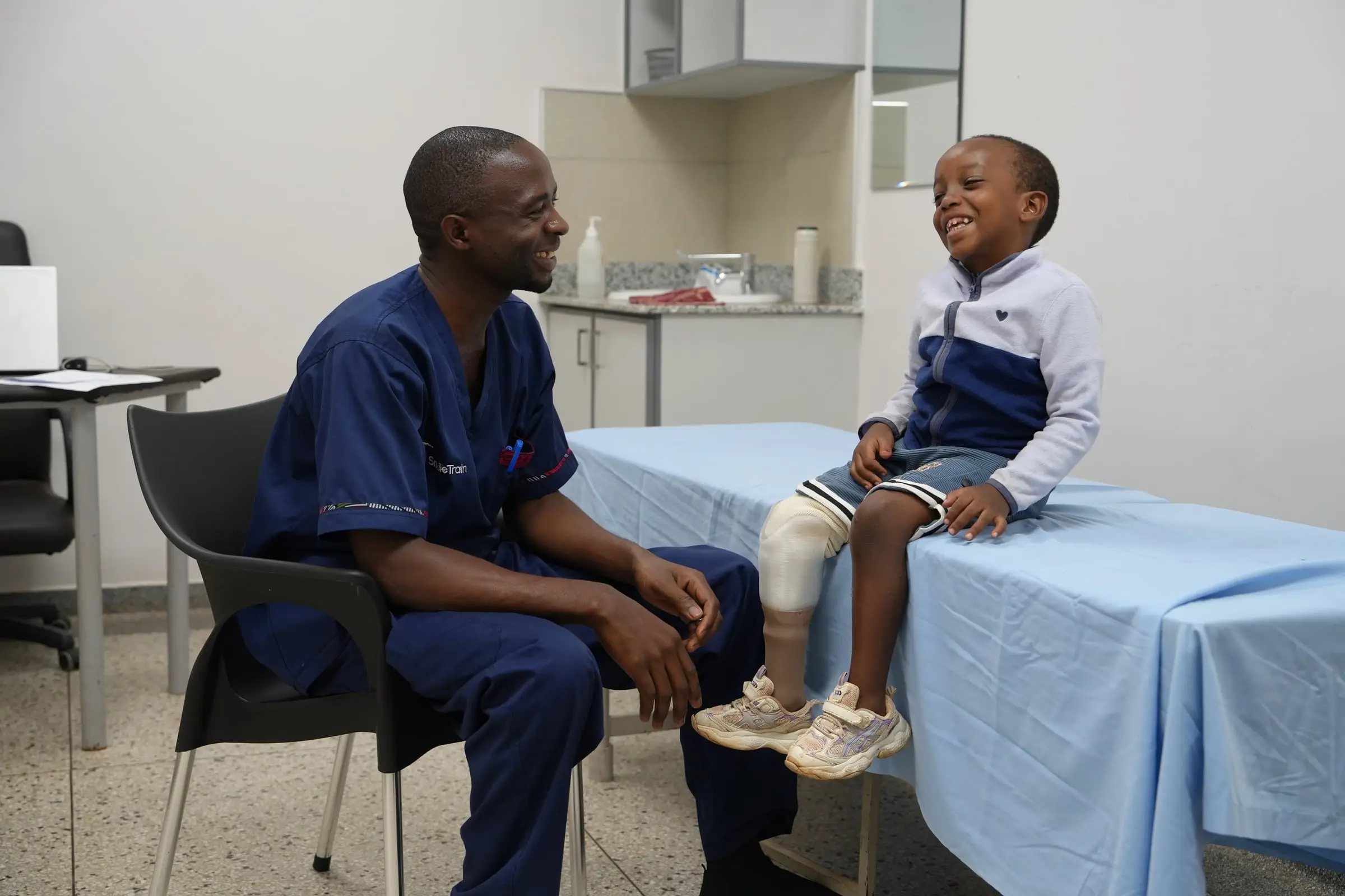 A clinician in navy scrubs and a young boy with a prosthetic leg smile at each other in a consulting room.