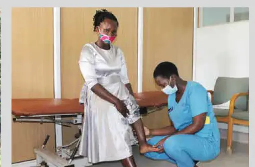 A young woman in a white dress and a pink face mask sits on a treatment couch while a clinician in blue scrubs kneels to examine her lower leg.