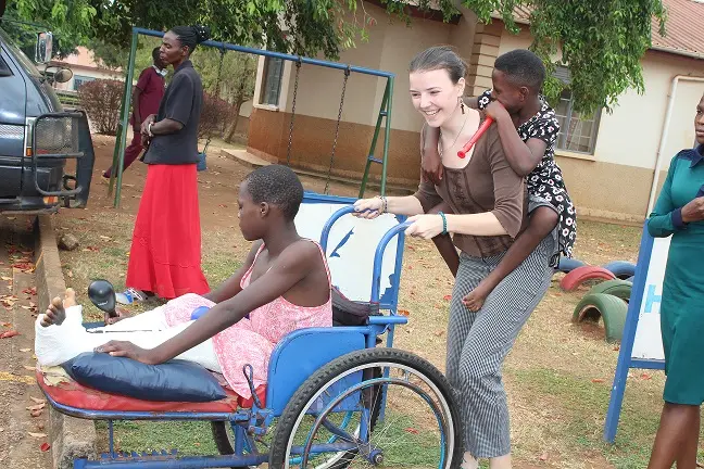 A young woman carrying a child on her back pushes a girl in a wheelchair past playground swings.