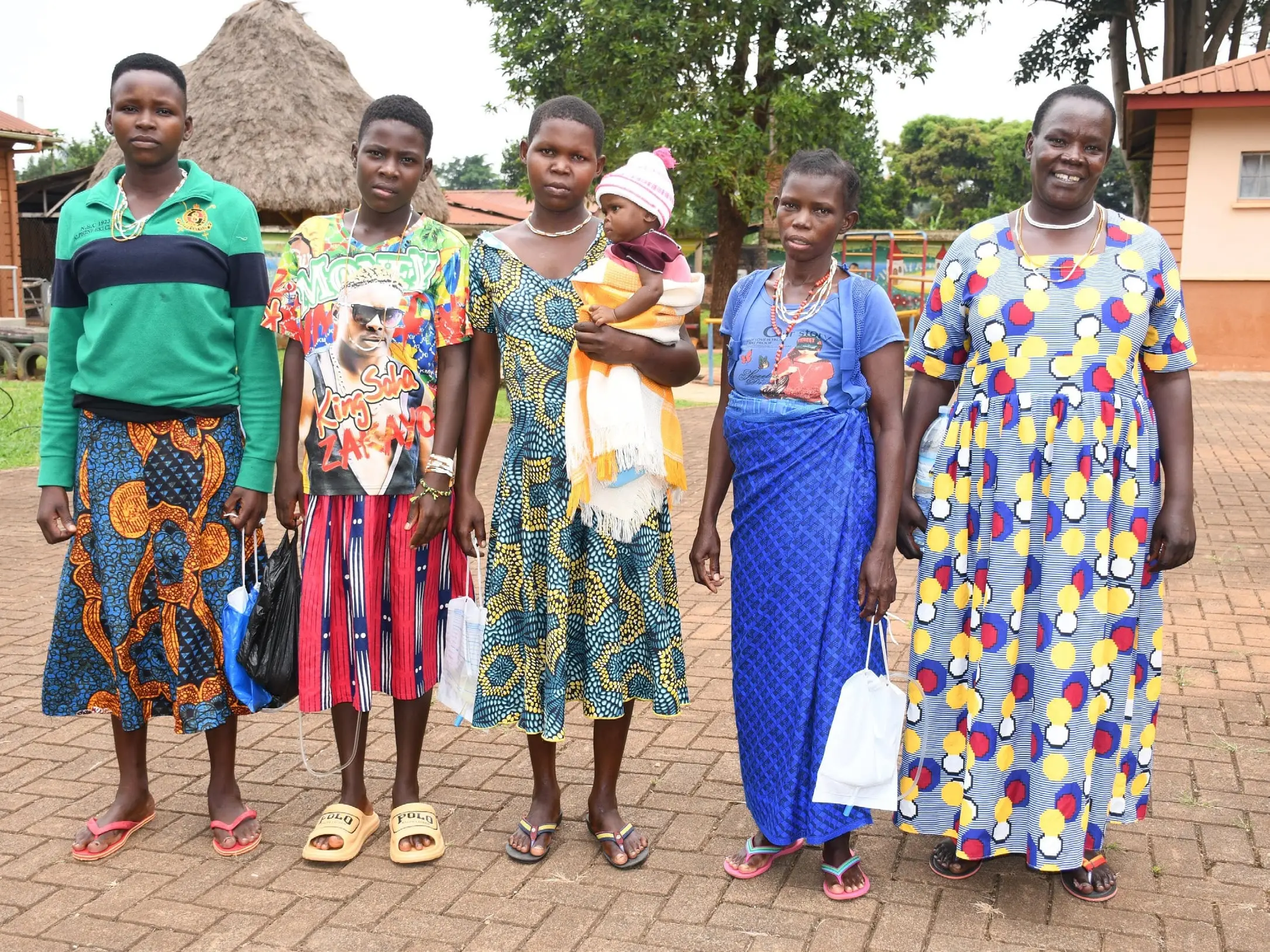 Six women in brightly patterned dresses, one holding a baby, stand together in the hospital grounds.
