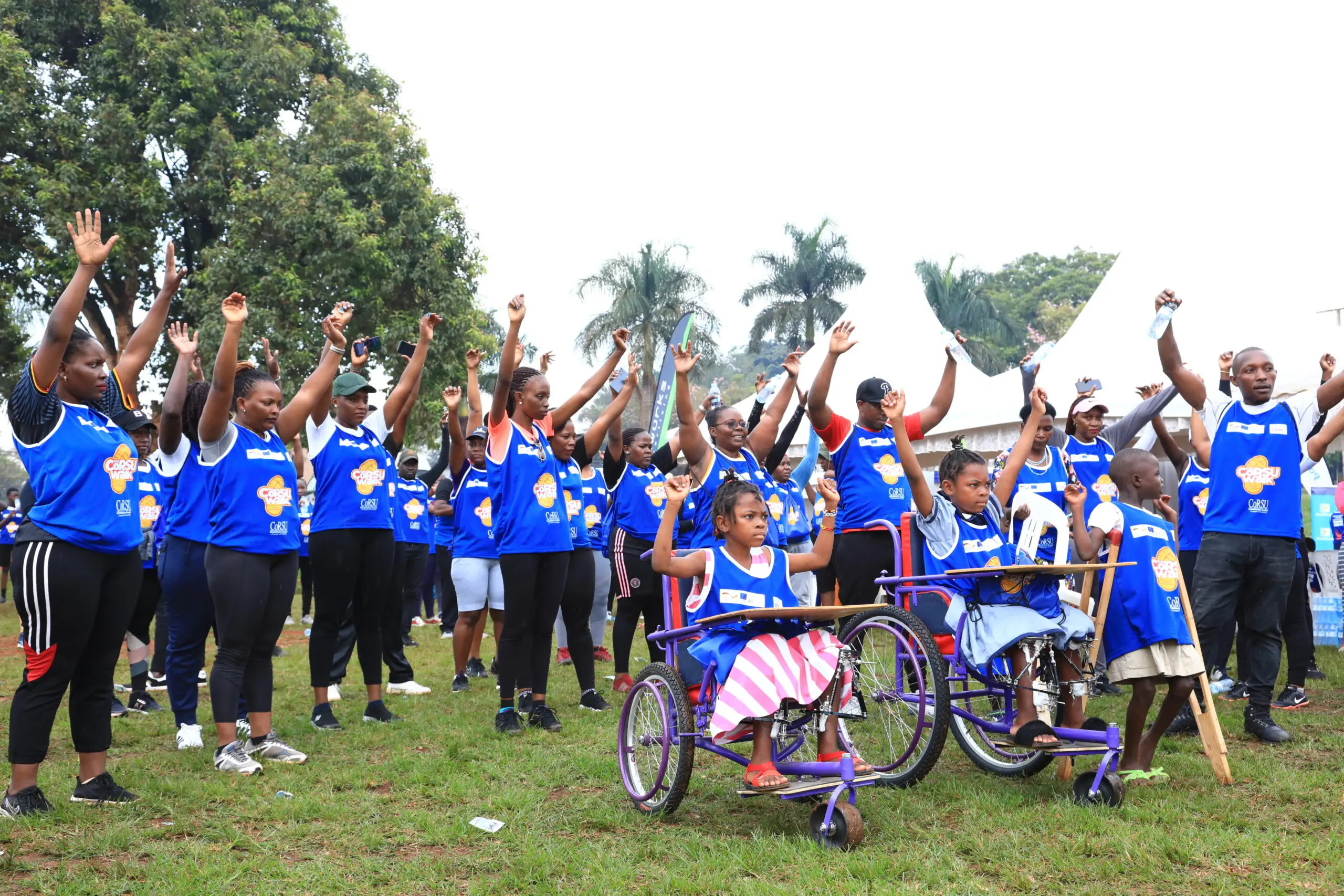 A crowd in blue CoRSU Walk bibs cheers with arms raised around children in wheelchairs and on crutches.
