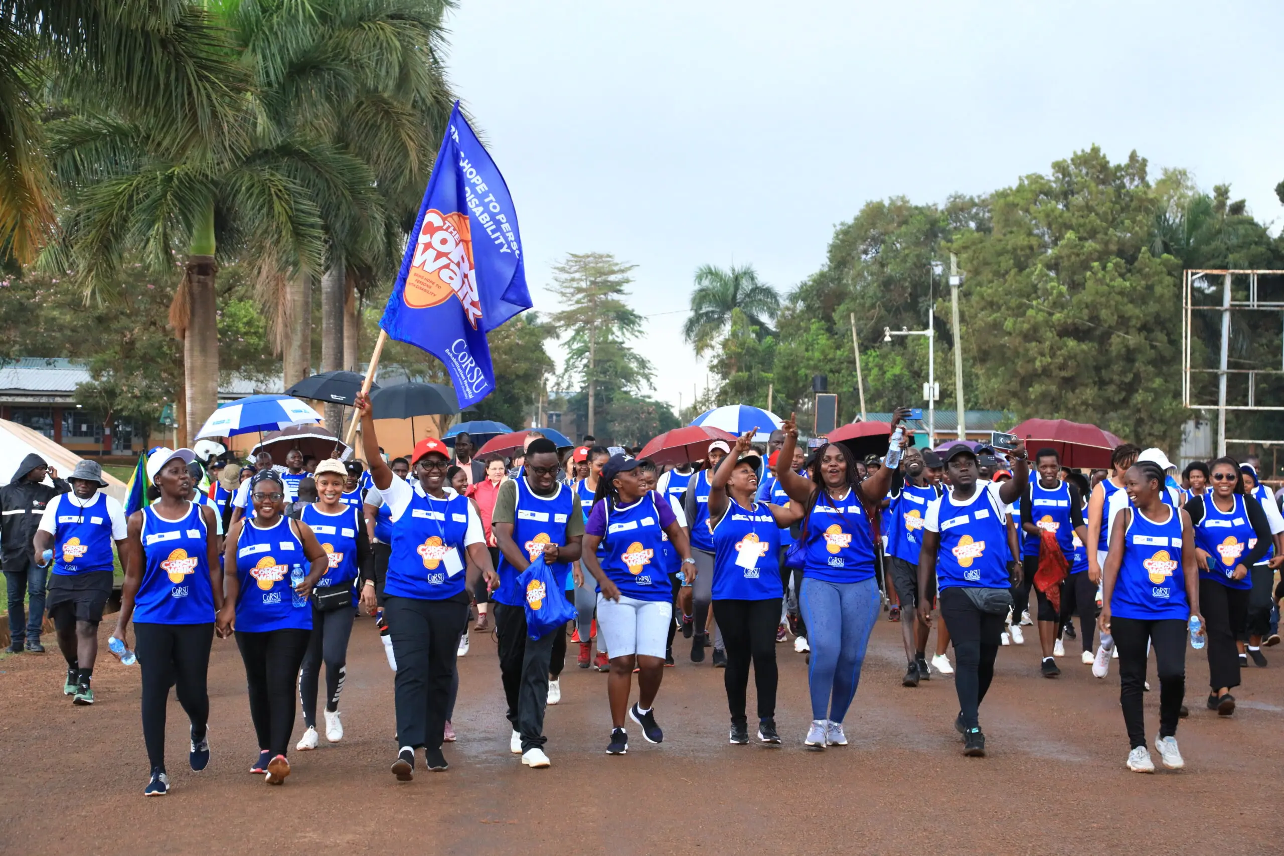 Walkers in blue bibs, some carrying umbrellas, follow a person holding a large CoRSU Walk flag.