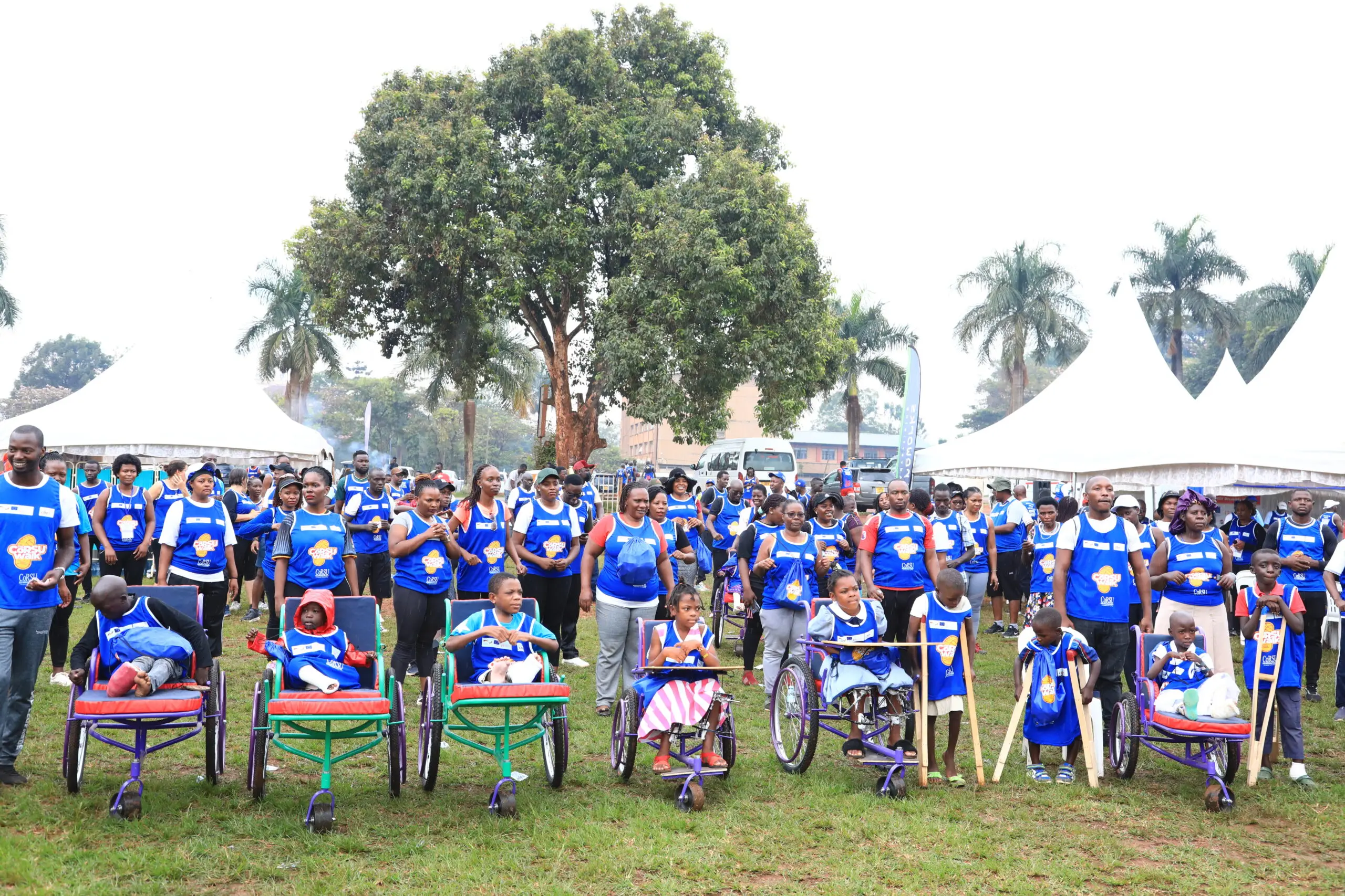 Hundreds of walk participants in blue bibs stand on a sports field behind a front row of children using wheelchairs, with white marquees behind them.