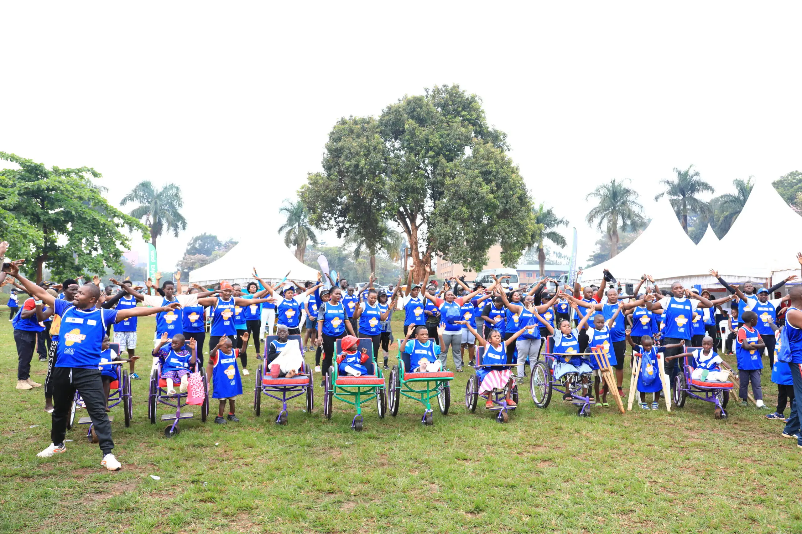 A large crowd of walkers in blue CoRSU Walk bibs wave from a sports field, with children in wheelchairs across the front row.
